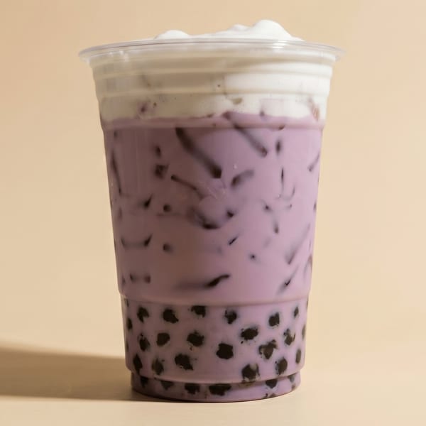 A clear plastic cup of purple taro bubble tea with dark boba pearls and a white foam topping.